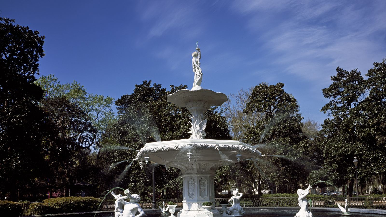 The white cast-iron fountain in Forsyth Park, Savannah, framed by trees under a blue sky